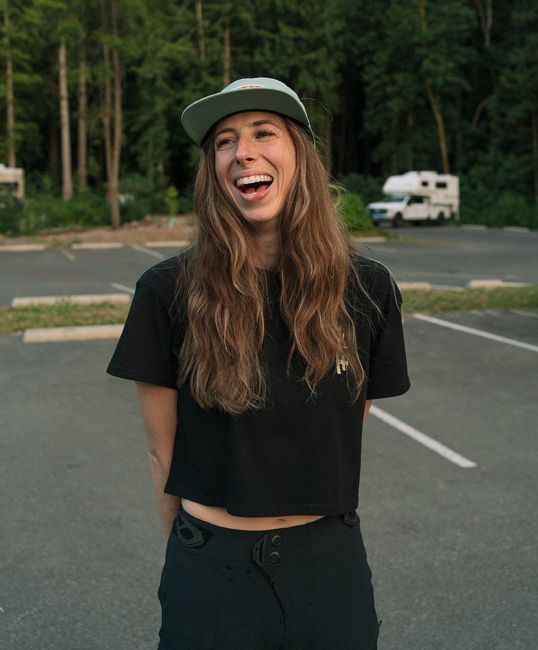Person with long hair wearing a black shirt and cap in a parking lot with trees in the background
