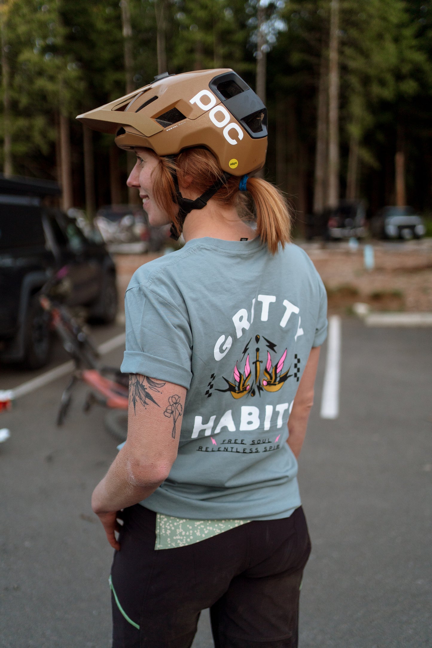 Person wearing a helmet and 'Gritty Habit' t-shirt in a parking lot with trees in the background