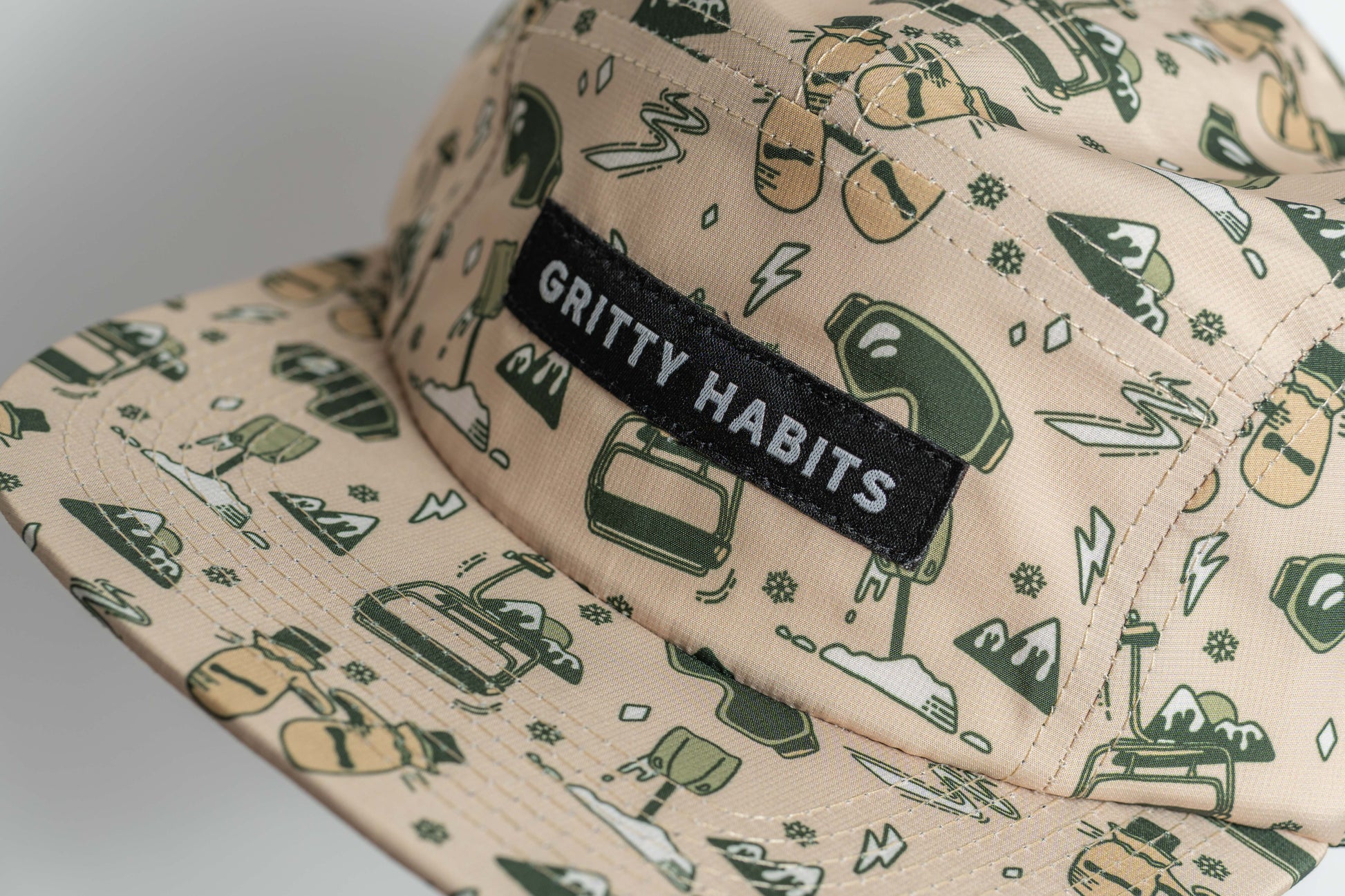 Beige cap with green ski patterns and 'Gritty Habits' label on a light gray background