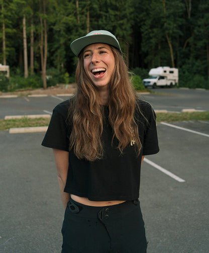 Person with long hair wearing a black shirt and cap in a parking lot with trees in the background
