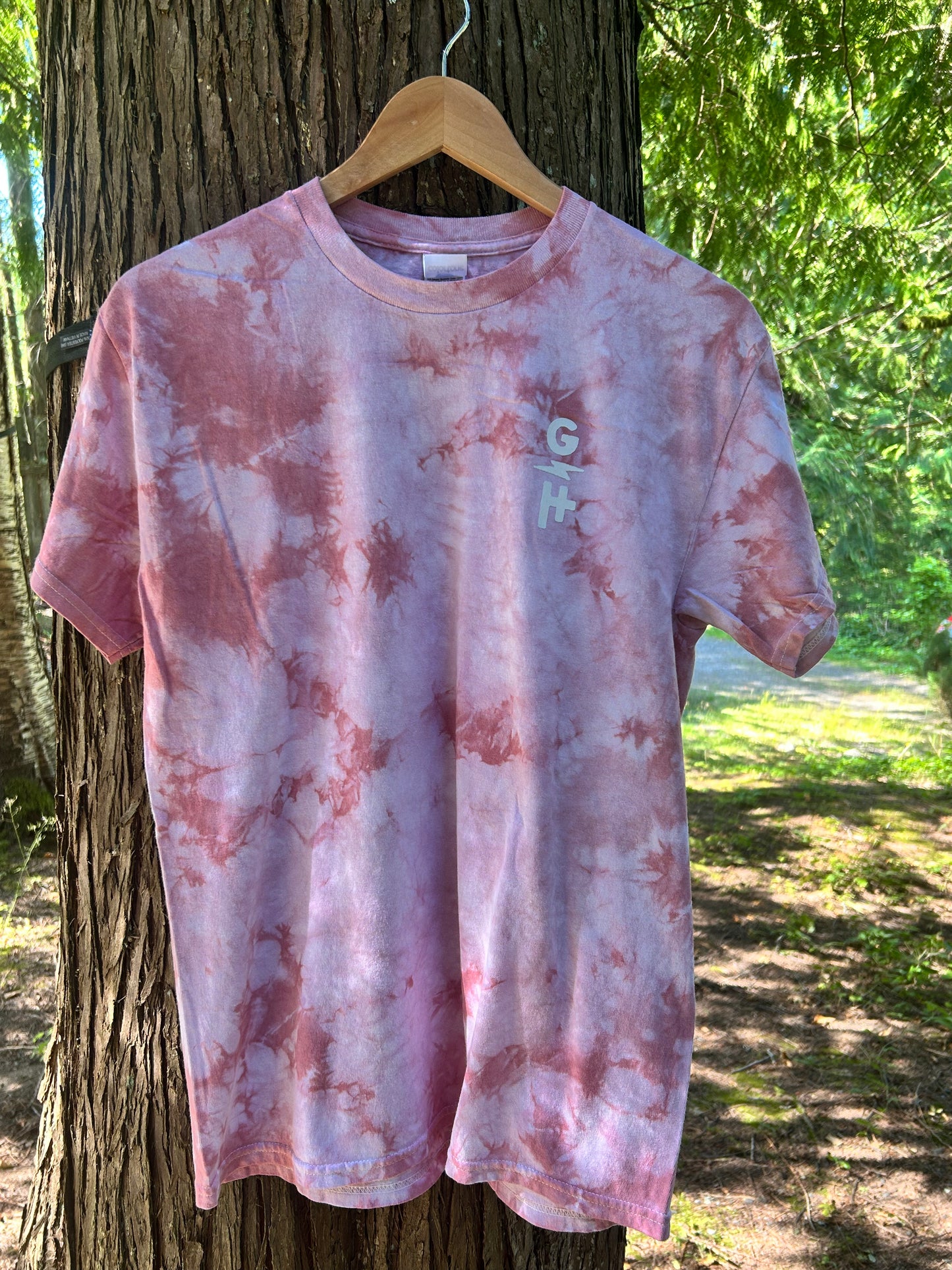 A pink tie-dye t-shirt with a small vertical 'GH' logo on the left chest, hanging on a wooden hanger from a tree outdoors.