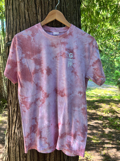 A pink tie-dye t-shirt with a small vertical 'GH' logo on the left chest, hanging on a wooden hanger from a tree outdoors.
