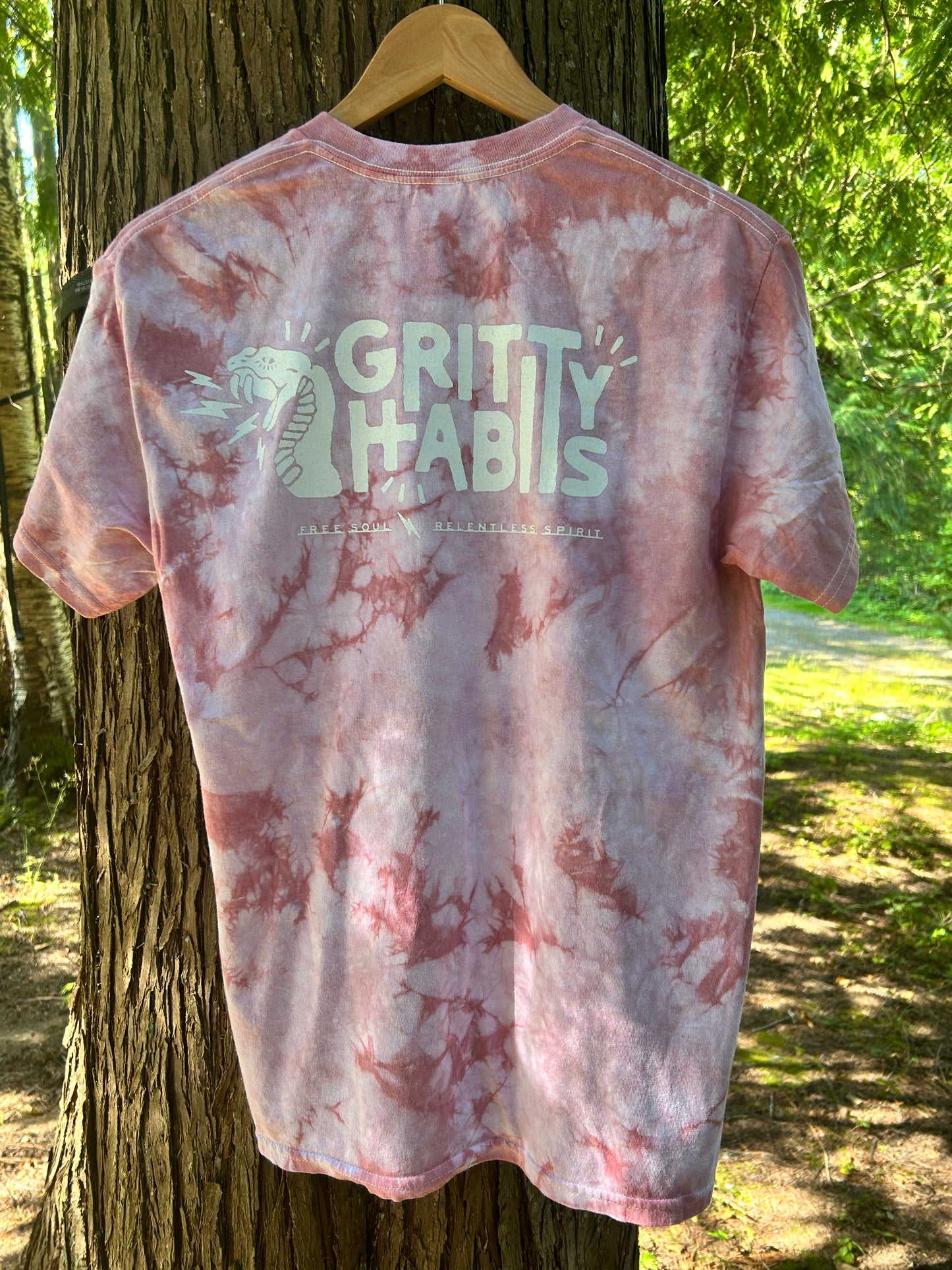 A short-sleeve tie-dye t-shirt with a pink and white marbled pattern, hanging on a wooden hanger from the back of a tree outdoors. The back of the shirt features a large white graphic that reads 'GRITTY HABITS' with a snake illustration and smaller text below.