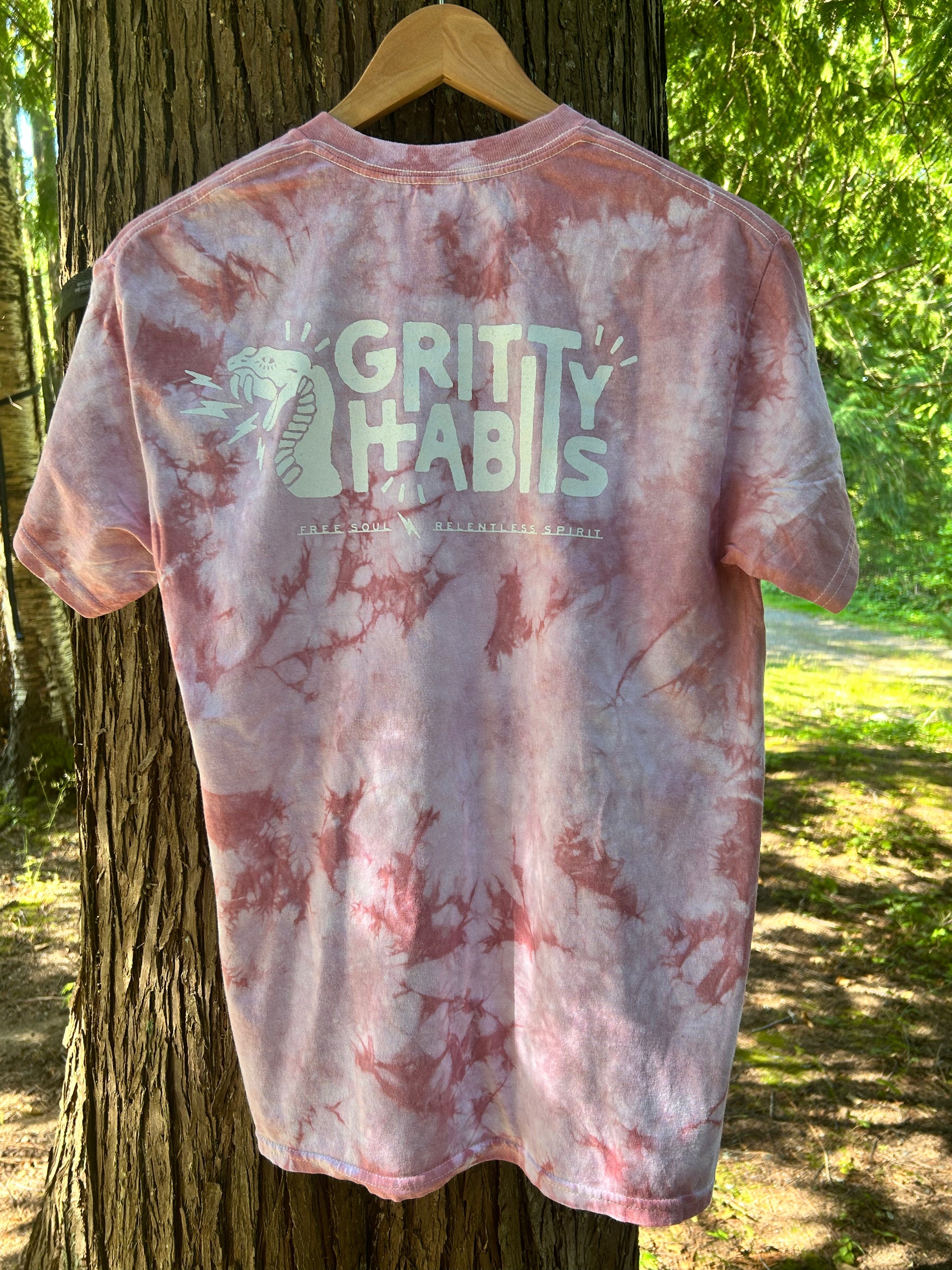 A short-sleeve tie-dye t-shirt with a pink and white marbled pattern, hanging on a wooden hanger from the back of a tree outdoors. The back of the shirt features a large white graphic that reads 'GRITTY HABITS' with a snake illustration and smaller text below.