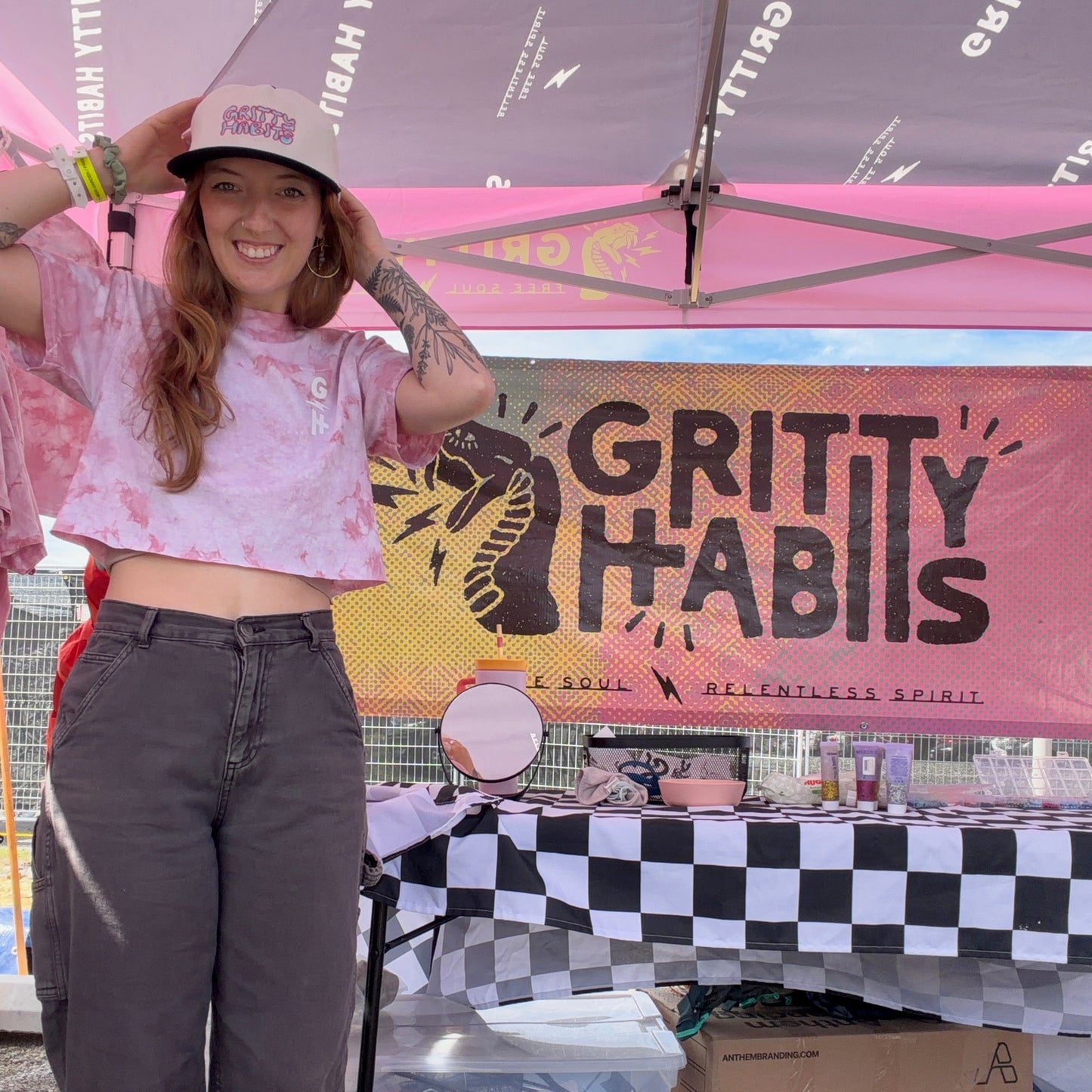 A woman stands under a pop-up canopy at an outdoor booth, wearing a pink tie-dye cropped T-shirt, dark gray jeans, and a beige cap with the Gritty Habits logo. A large Gritty Habits banner hangs behind her above a table with a black-and-white checkered tablecloth and a cardboard box labeled Anthem Branding.