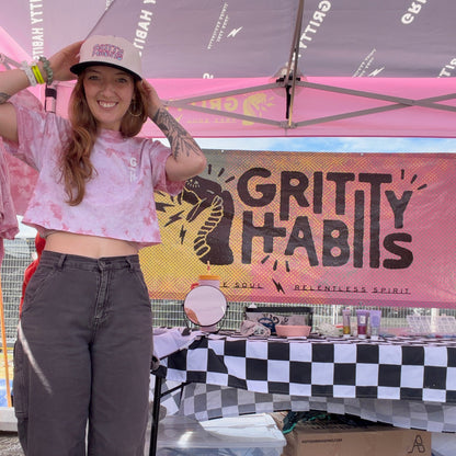 A woman stands under a pop-up canopy at an outdoor booth, wearing a pink tie-dye cropped T-shirt, dark gray jeans, and a beige cap with the Gritty Habits logo. A large Gritty Habits banner hangs behind her above a table with a black-and-white checkered tablecloth and a cardboard box labeled Anthem Branding.